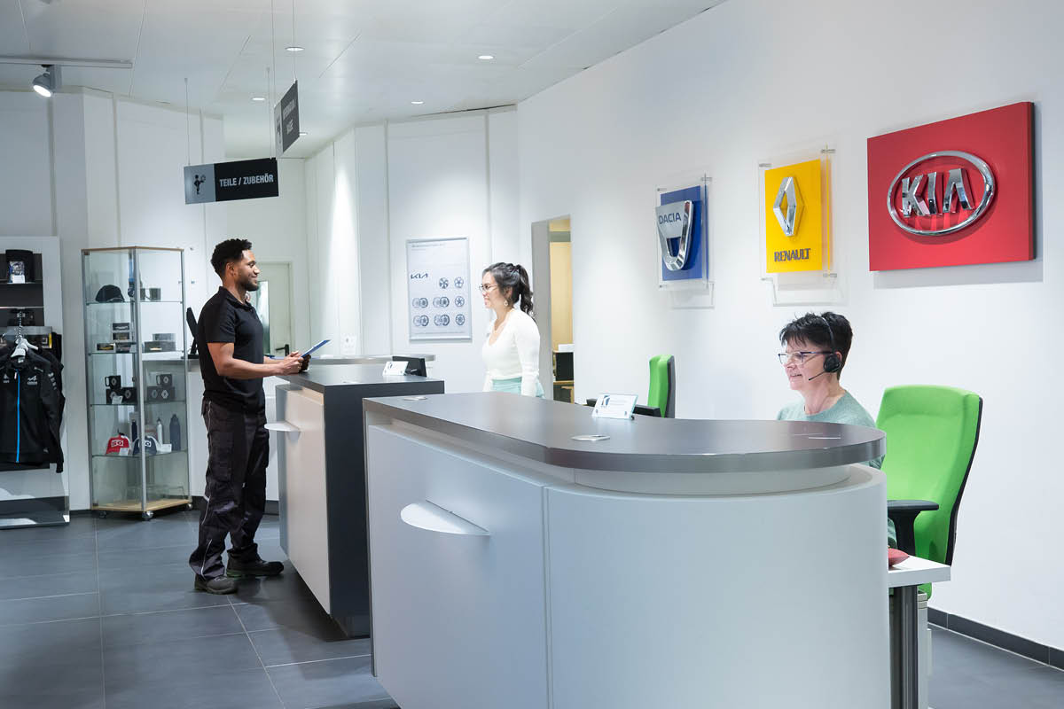 Empfangsbereich eines Autohauses. Mehrere Personen stehen oder sitzen an einer modernen Theke.