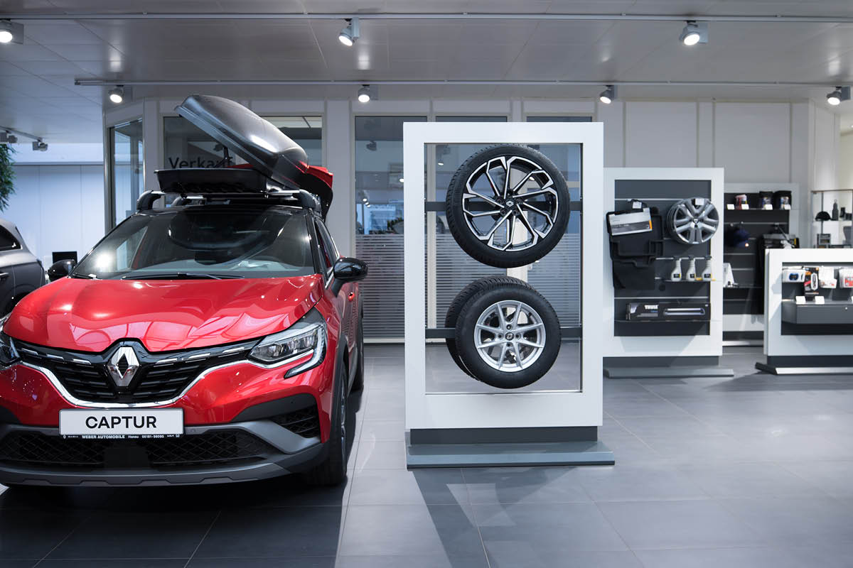 Ausstellungsraum des Autohauses. Im Vordergrund steht ein roter Renault Captur mit einer geöffneten Dachbox. Rechts davon sind zwei ausgestellte Autoreifen und Felgen, dahinter Zubehörteile und weitere Produkte an Regalen.