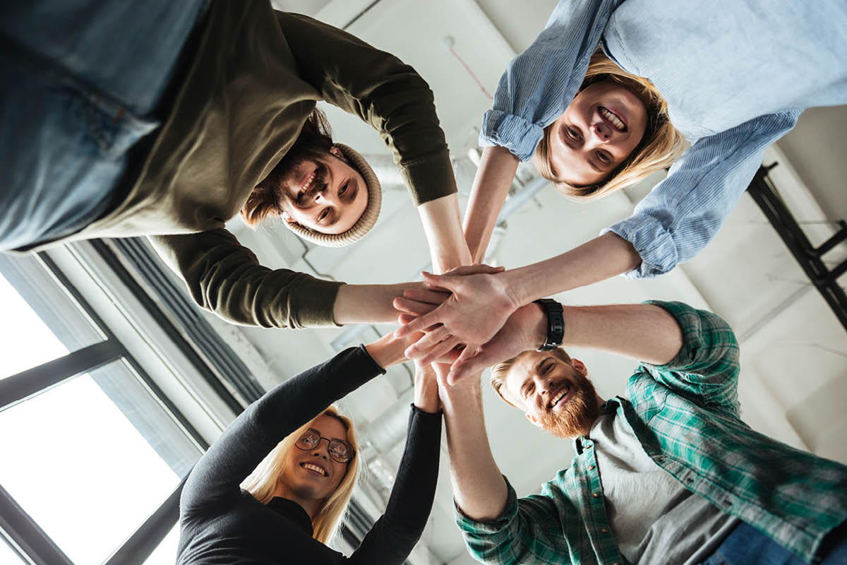 Eine Gruppe von Menschen, die ihre Hände in der Mitte übereinanderlegen. Die Perspektive ist von unten aufgenommen.