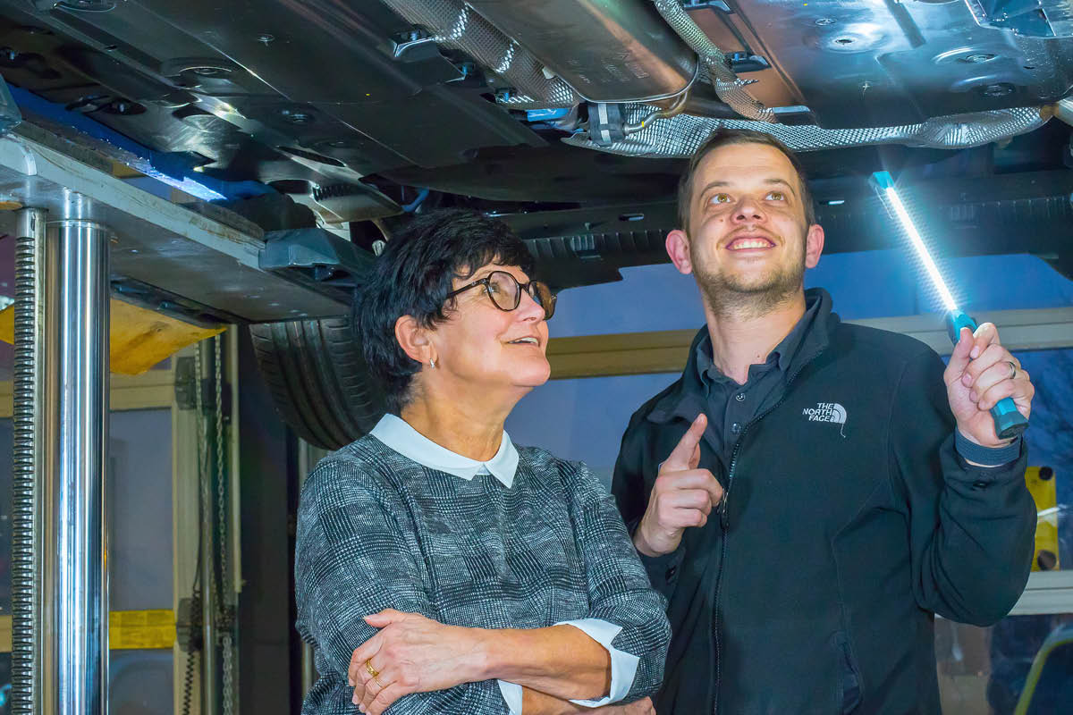 Zwei Personen stehen unter einem aufgebockten Auto in einer Werkstatt. Eine Person hält eine Lampe und zeigt auf einen Bereich unter dem Fahrzeug, während die andere aufmerksam zuhört.